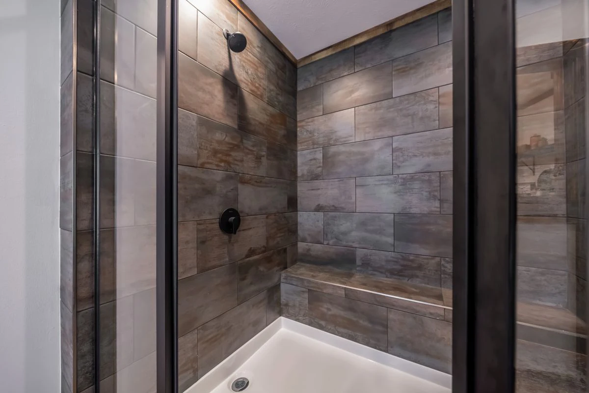 2c4c4e46-d898-45fb-b28e-10d80cfcb127 A walk-in shower with large, rectangular, dark wood-look tiles, a built-in bench, a black showerhead, and a white shower pan.