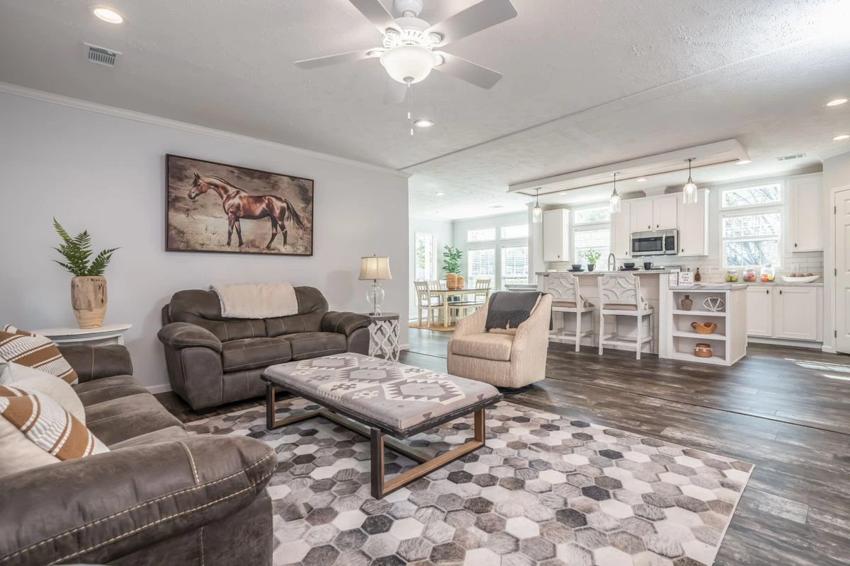 A modern living room with brown sofas, an armchair, a patterned rug, a coffee table, and a horse painting on the wall, adjacent to a white kitchen and dining area.