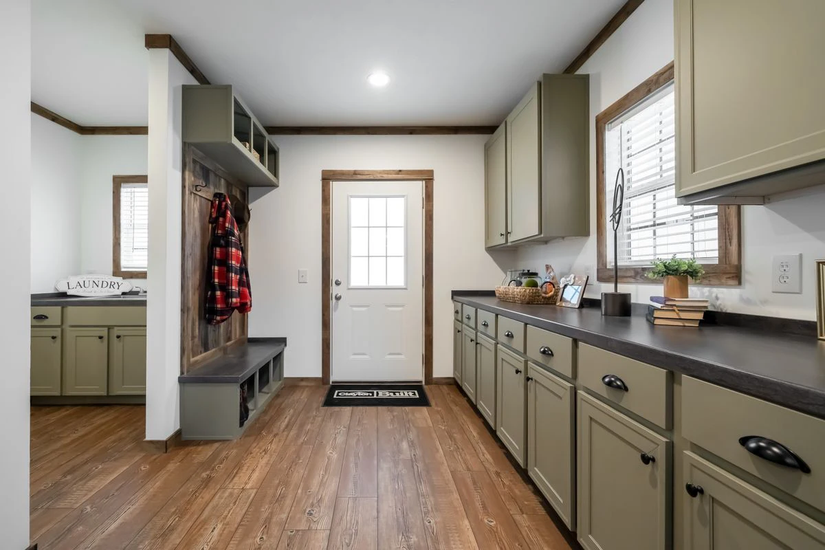 42357513-41e6-4ce9-b5c1-7eaf33dc172a A laundry and mudroom area with green cabinets, a white back door, wooden floors, shelves, a plaid jacket hanging, and laundry supplies on the counter.