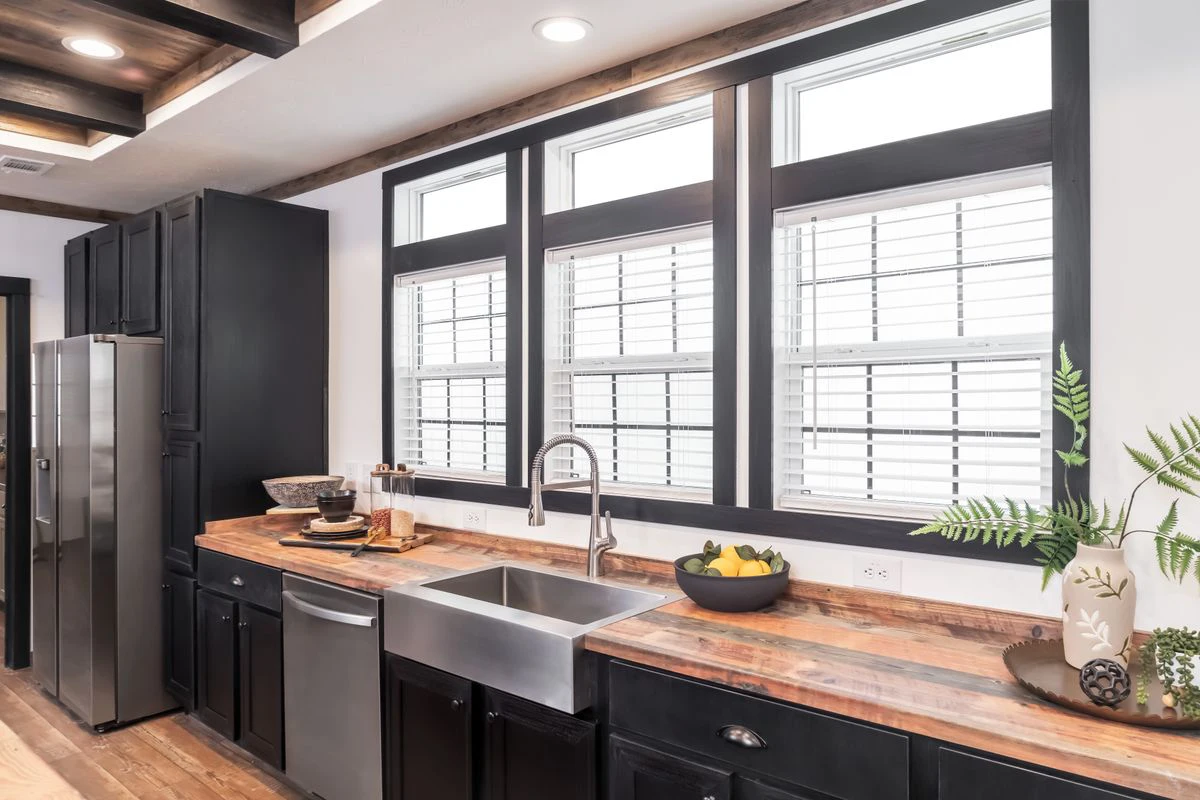 42366931-0c14-4f18-91ae-9eede5eb6c4d Modern kitchen with black cabinets, stainless steel appliances, wood countertops, a large farmhouse sink, three windows with white blinds, and decorative plants and bowls on the counter.