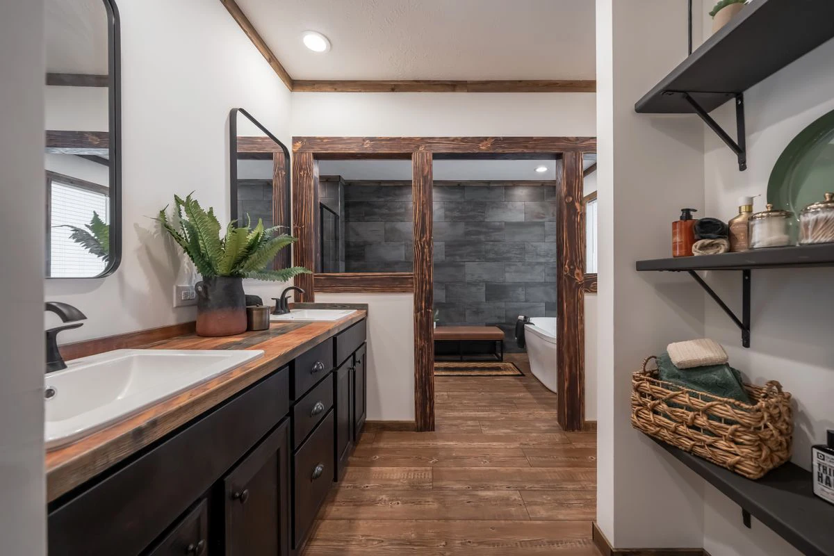 4af6c039-efa7-44e6-bf3a-b68fbbb34a16 Modern bathroom with double sinks, wood countertops, dark cabinets, wall shelves, potted plant, and a view of a bathtub and tiled shower area in the background.