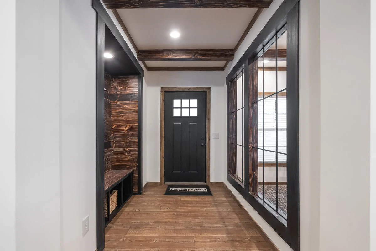 4b960246-6c5b-48a9-848c-a1f8756f7e76 A hallway with a black front door, wood flooring, wooden bench with storage on the left, and large windows on the right. Ceiling beams and white walls are visible.
