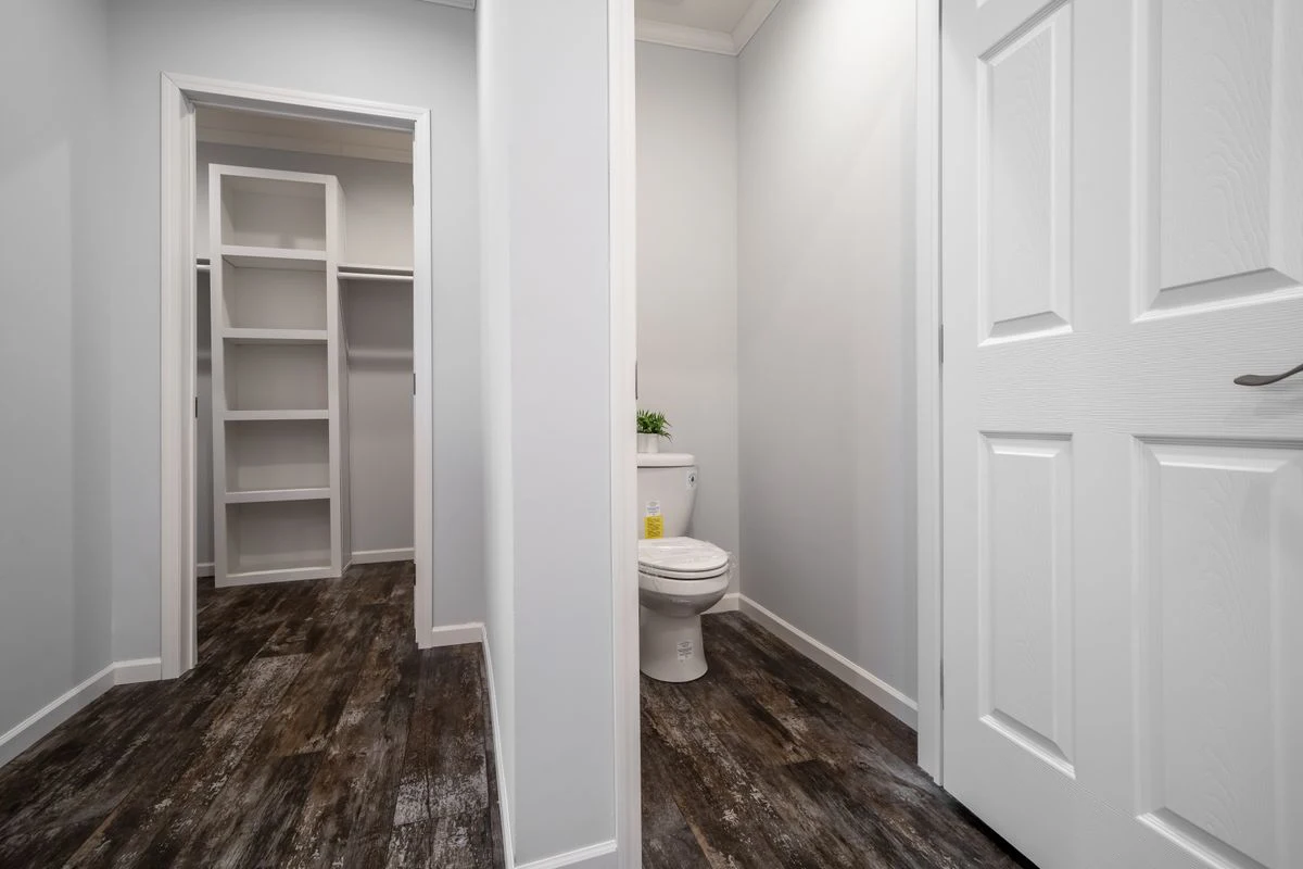 A small bathroom with a white toilet, gray walls, and dark wood flooring, next to an open doorway leading to a walk-in closet with built-in shelves.