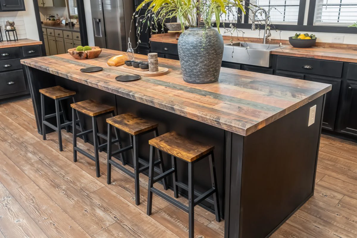 6070df3b-b1a5-4f57-9398-bffae8784f7f A large kitchen island with a wood countertop, four wooden stools, a gray vase with greenery, and kitchen items on top. Dark cabinets and wood flooring surround the space.