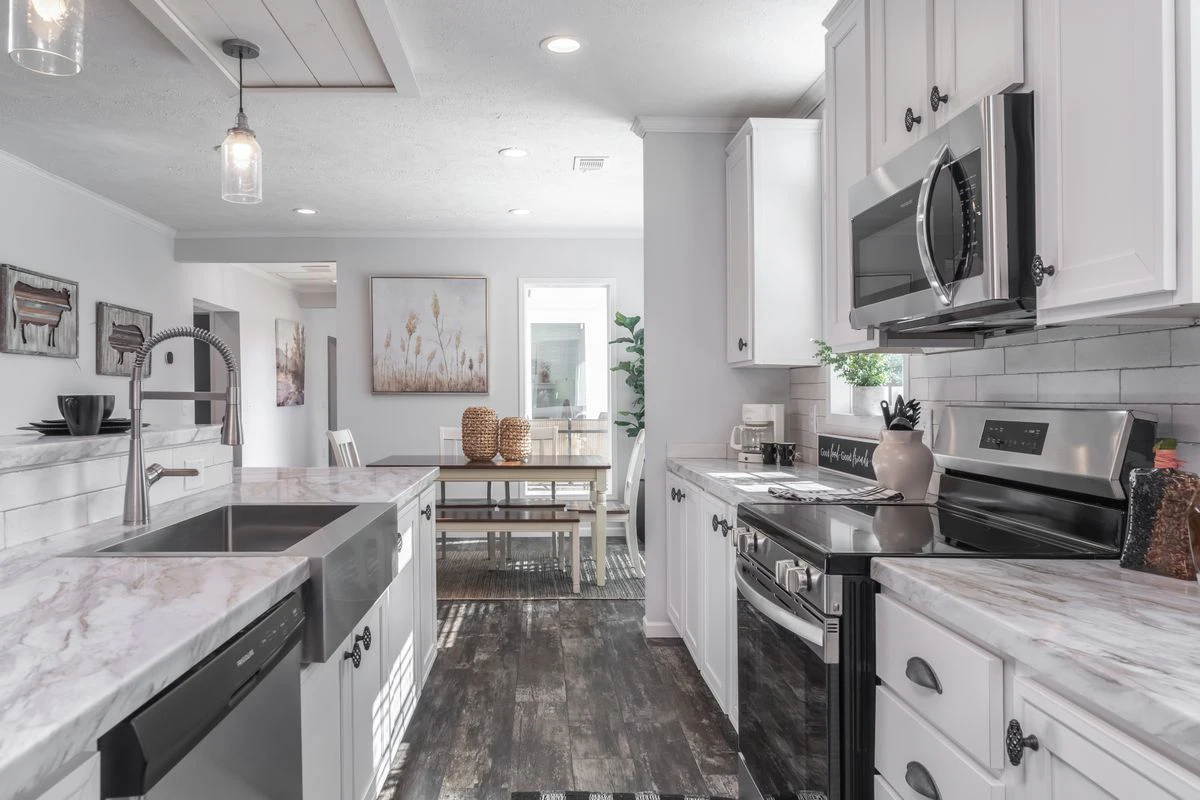 Modern kitchen with white cabinets, marble countertops, stainless steel appliances, and a farmhouse sink, opening to a dining area with a table and chairs.