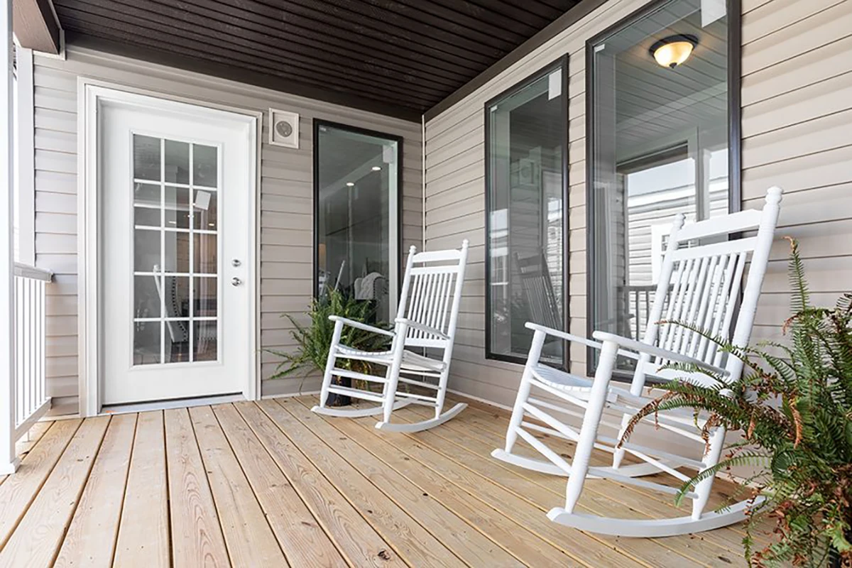 A porch with light wood flooring, two white rocking chairs, potted ferns, large windows, and a glass door under a dark ceiling.