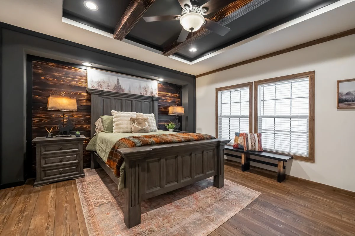72f2712d-da3b-4068-bbfd-5c0493c237a1 A bedroom with a wooden bed, nightstands, lamps, a bench by two windows, a ceiling fan, and a wood-accented wall with framed art above the bed.
