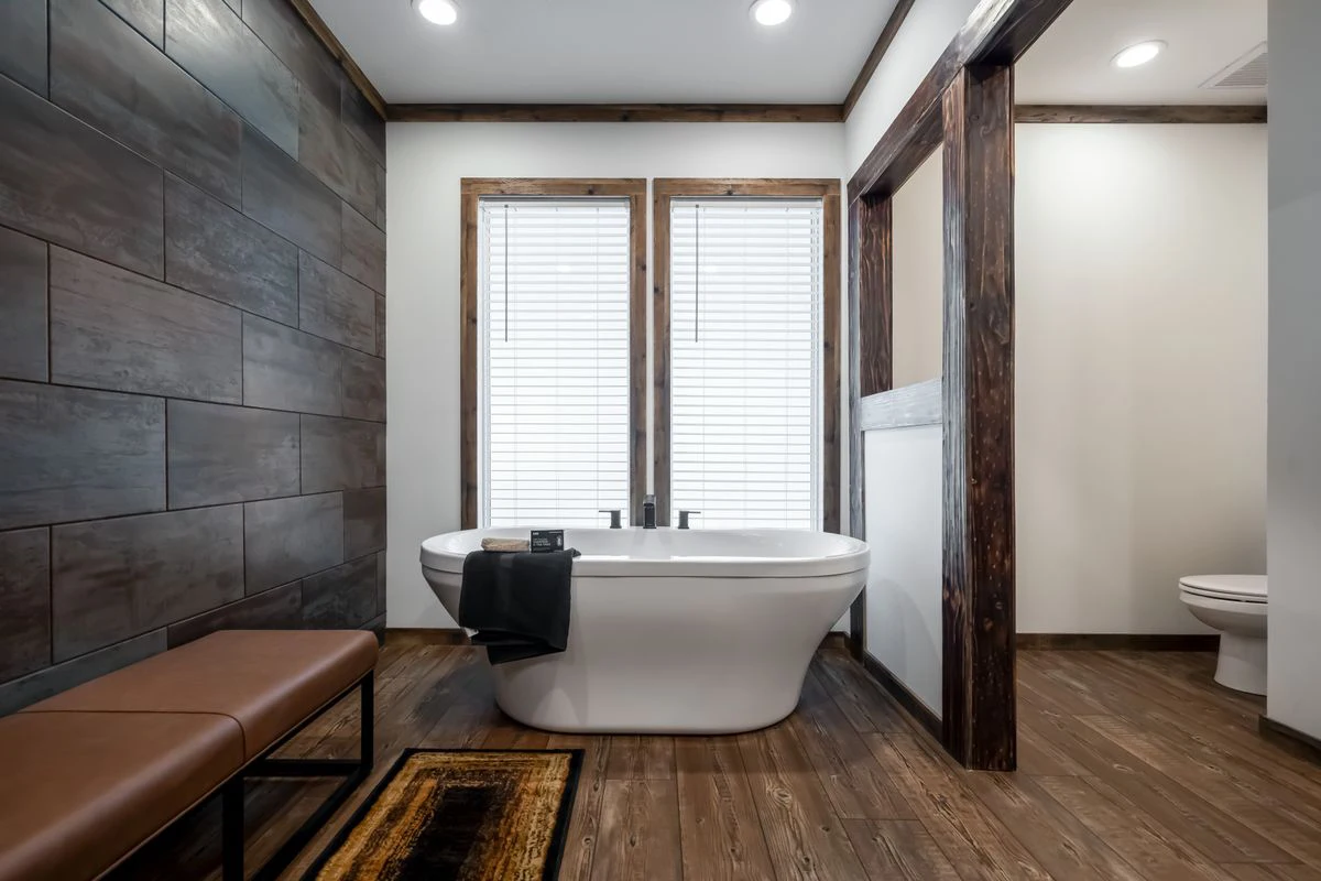 b1031b58-d4ef-4aad-840f-845990c5cfd0 Modern bathroom with a white freestanding tub, wood floors, dark tile wall, bench, two large windows with blinds, and a toilet partially visible in a separate nook.