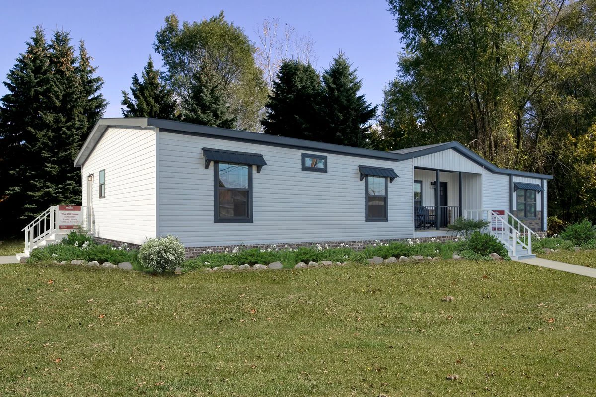 bfb8617a-086e-488a-9572-bbfe80c674f3 A single-story manufactured home with white siding, black trim, and a front porch, surrounded by grass and trees in the background.