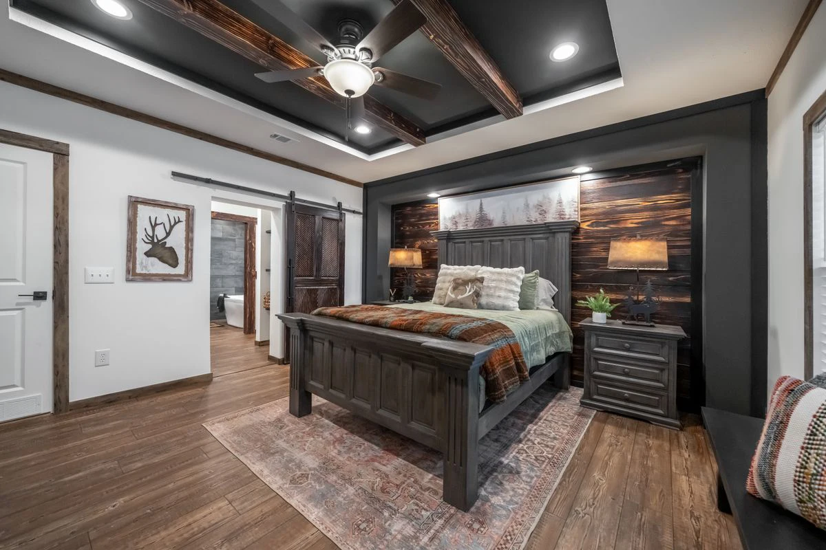 d6a10b34-9434-49df-84dd-677c596db044 A rustic bedroom with a dark wooden bed, matching nightstands, barn door, ceiling fan, lamps, and nature-themed decor, featuring wood floors and a patterned rug.