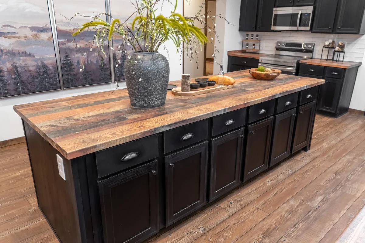 db33645a-bce9-4f1f-abad-c8c8e0006a63 A kitchen with a large island featuring dark cabinets and a wood countertop, decorated with a vase of greenery and various kitchen items.