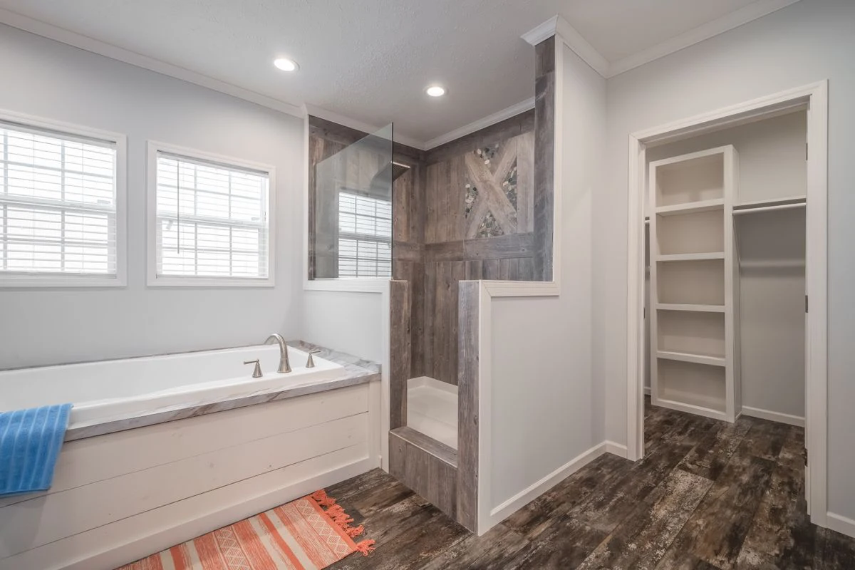 Modern bathroom with a white bathtub, walk-in shower with gray tiles, wooden floors, recessed lighting, and an open walk-in closet with shelves.