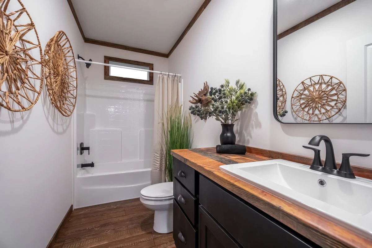 fca434b2-6714-4e2e-be32-3f84f1506e1d A bathroom with a white bathtub and shower, a toilet, dark wood vanity with a white sink, black fixtures, and decorative wall art and plants.
