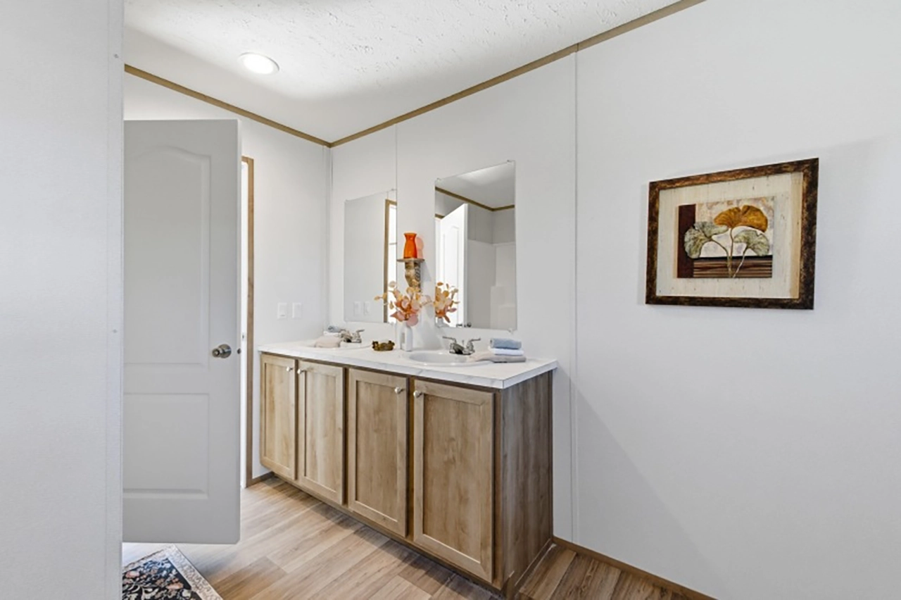 A bathroom with double sinks, wooden cabinets, two mirrors, a decorative lamp, and a framed floral picture on a white wall.