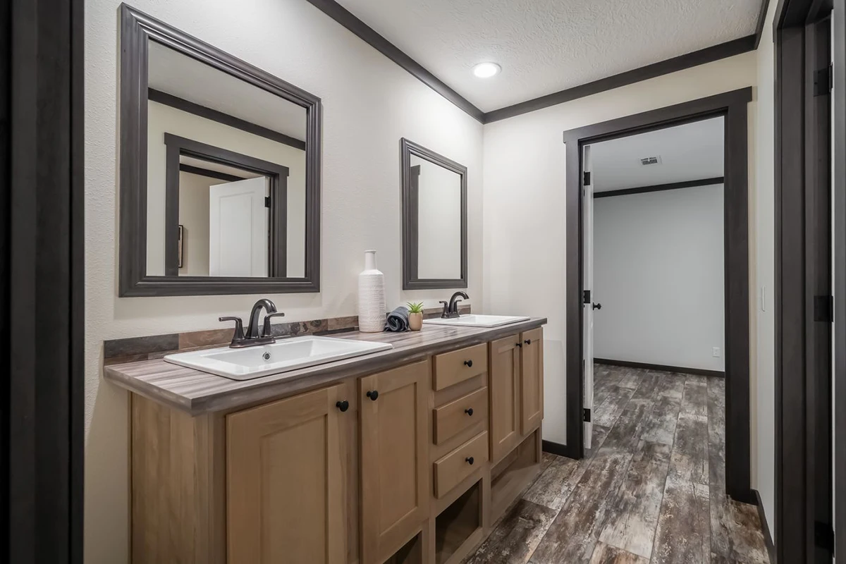10891fb6-e400-4d27-975a-dddd4319ae79 Modern bathroom with double sinks, wooden vanity, two mirrors, a plant, and a soap dispenser. Doorway leads to a room with wood-patterned flooring and white walls.