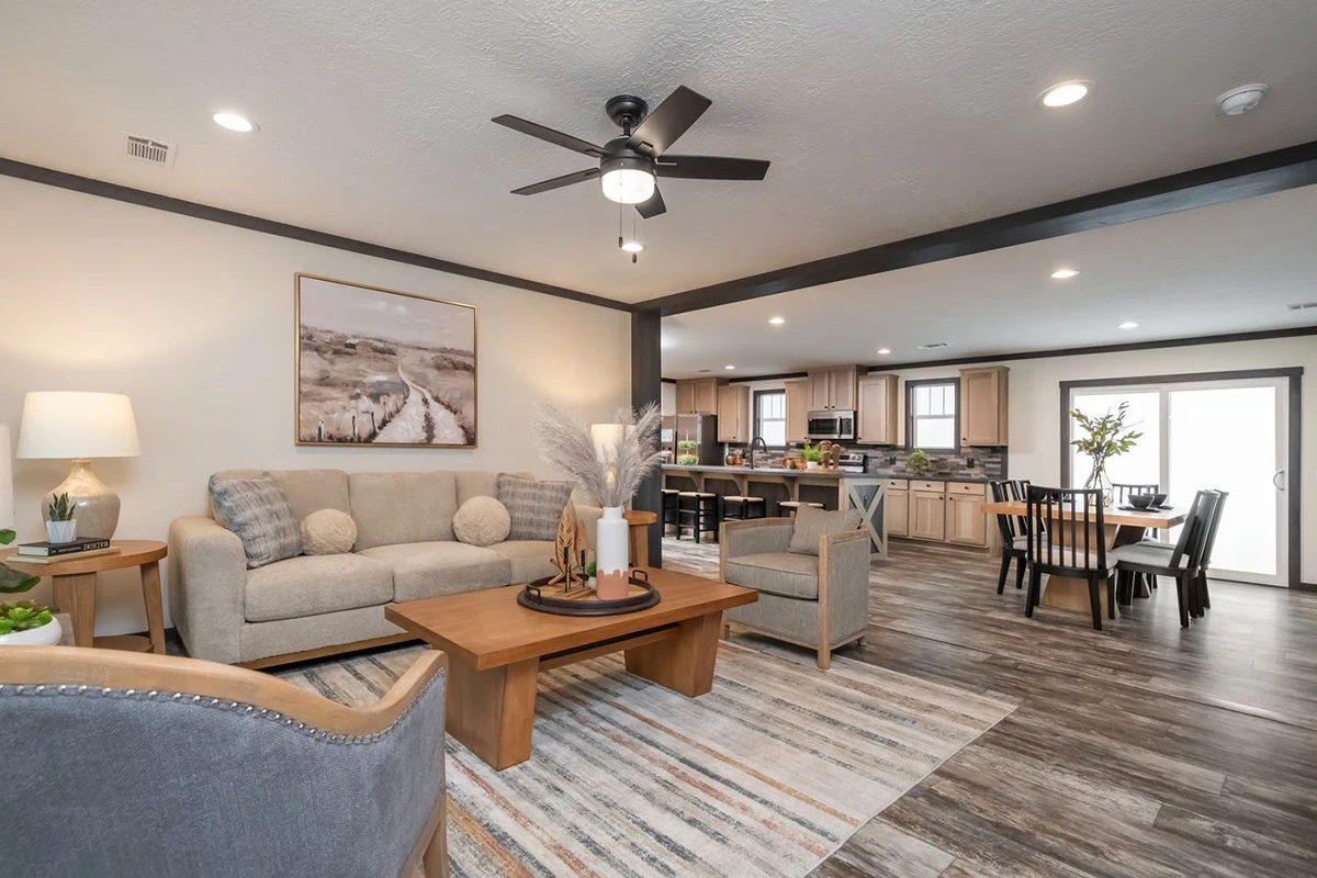 11899ab1-795d-4ef9-9fd0-083189b0adbf Open-concept living room and kitchen with neutral decor, modern furniture, wooden tables, area rug, and dining area visible in the background.