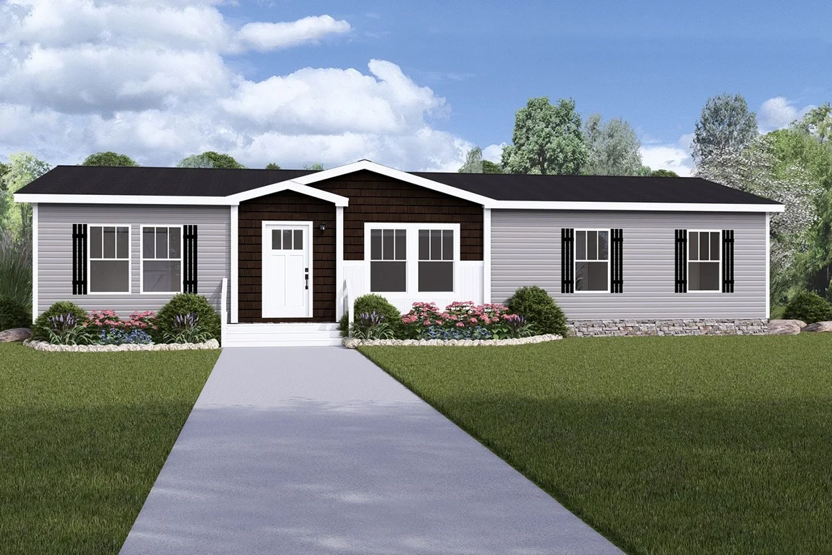 A single-story manufactured home with gray siding, black shutters, white trim, landscaped shrubs, and a concrete walkway leading to the front door, set against a backdrop of trees and blue sky.
