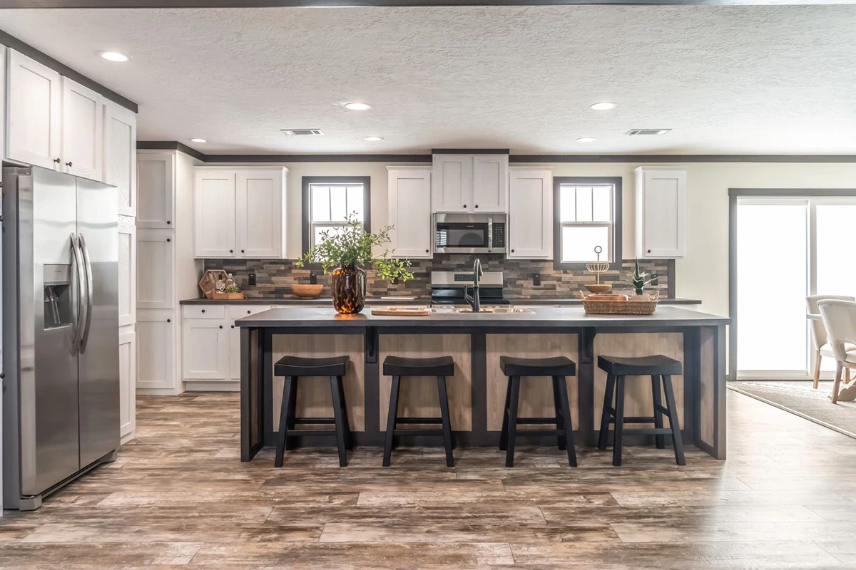Modern kitchen with white cabinets, stainless steel appliances, a large island with four black stools, and wood-style flooring. Natural light comes in from multiple windows.