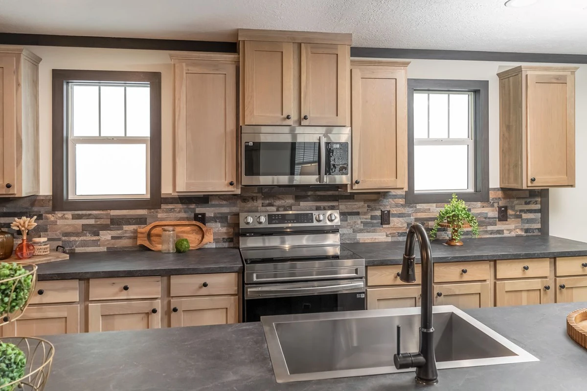 5965ac68-7b13-458b-863b-b2781a71ada5 Modern kitchen with light wood cabinets, stainless steel appliances, stone backsplash, and a black sink faucet centered in a dark countertop island. Two windows provide natural light.