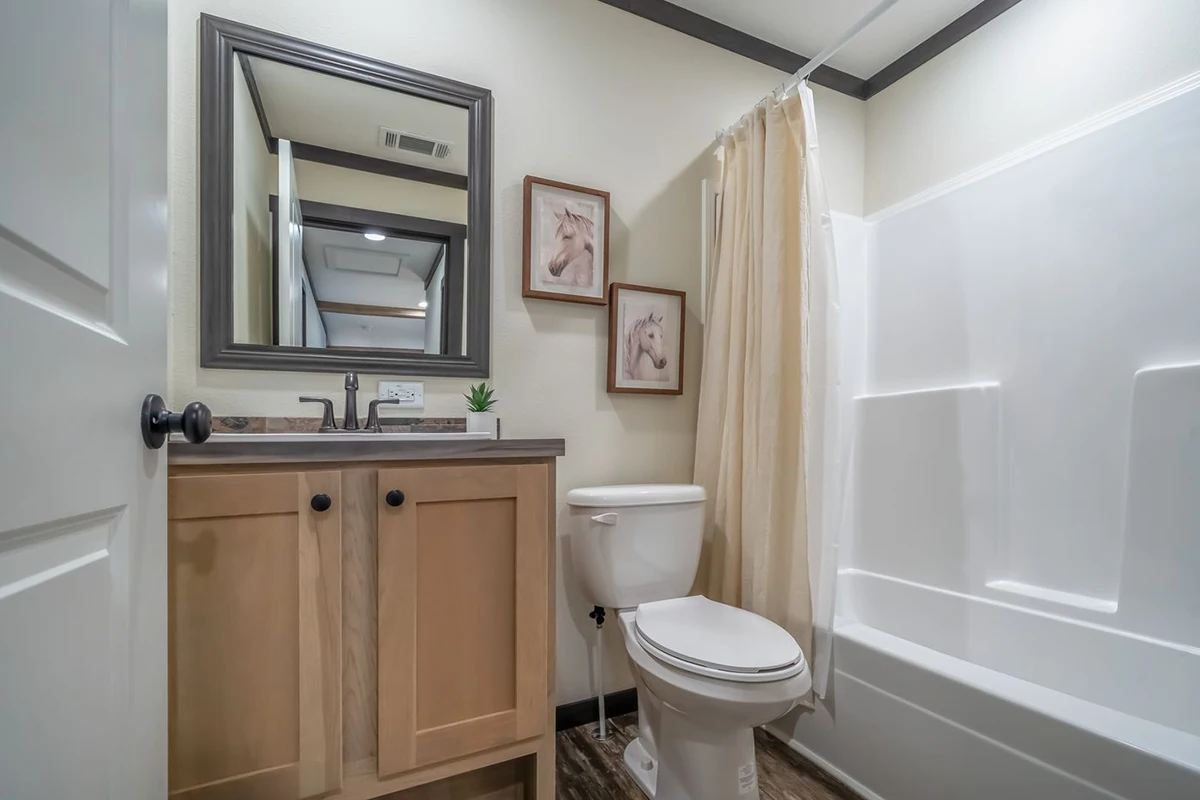 68a44164-90c8-4e6e-842f-6c964c28c3c3 A small bathroom with a wood vanity, sink, mirror, toilet, bathtub with a shower curtain, and two horse pictures on the wall.