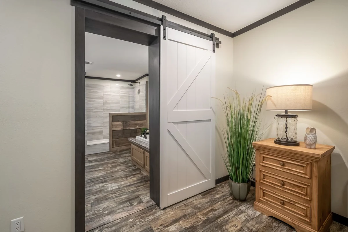 7a5a411e-e7fe-4e4d-a12b-ee08201bbf98 A bathroom with a bathtub is visible through a partially open sliding barn door; a lamp, tall plant, and wooden dresser sit in the foreground on wood flooring.