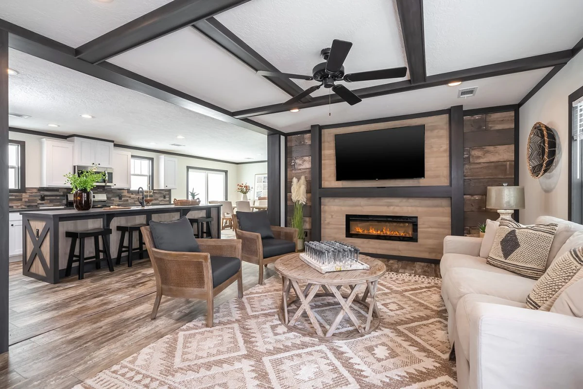 Modern open-concept living room and kitchen with wooden accents, a ceiling fan, wall-mounted TV, electric fireplace, sofa, armchairs, and a patterned rug.