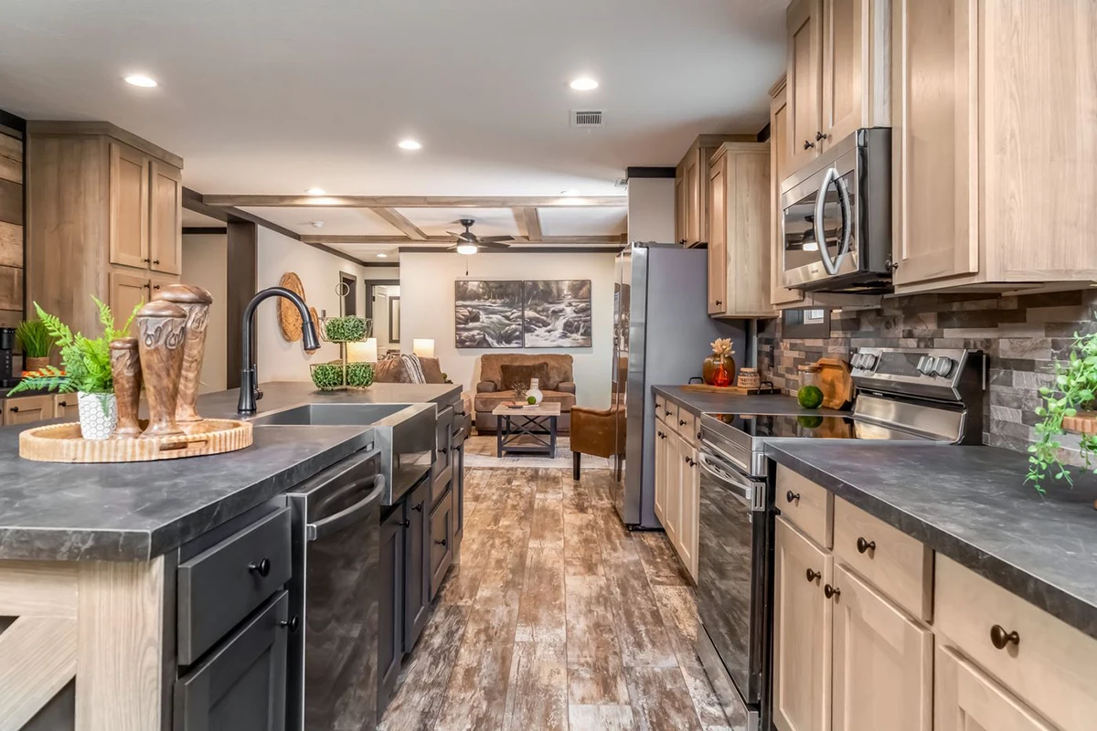 86142122-e7ff-4bd1-a3ef-6d85d5d2a3da Modern kitchen with wood cabinets, black countertops, stainless steel appliances, a double sink, and an open view into a living room with seating and decor.