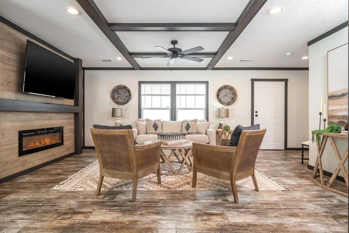 Modern living room with wood flooring, neutral furniture, a wall-mounted TV, electric fireplace, and two windows behind a beige sofa. Two chairs and a round coffee table are centered on a rug.