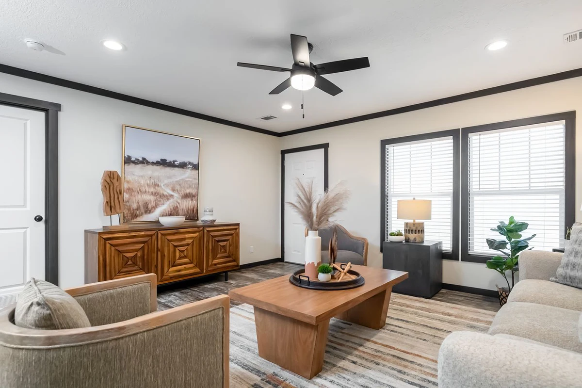 8e47d896-bc52-4796-8752-a4389d0c70e9 Modern living room with a beige sofa, wooden coffee table, sideboard, neutral decor, large windows with blinds, ceiling fan, and framed landscape art on the wall.