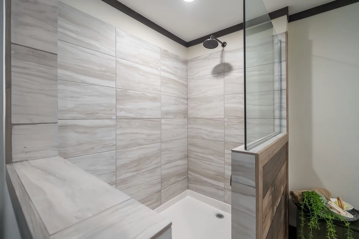 9d82ffd4-f3df-414b-9712-f96bb20251a0 Modern walk-in shower with light gray marble-patterned tiles, a black overhead showerhead, and a partial glass divider. A small corner with a plant and toiletries is visible outside the shower.