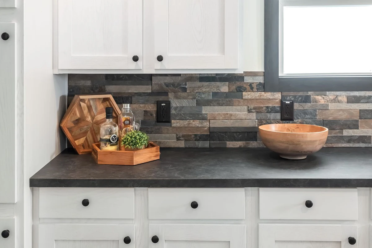 A kitchen countertop with a wooden bowl, a wooden tray holding bottles and a small plant, and a stone tile backsplash. White cabinets with black knobs are below the counter.