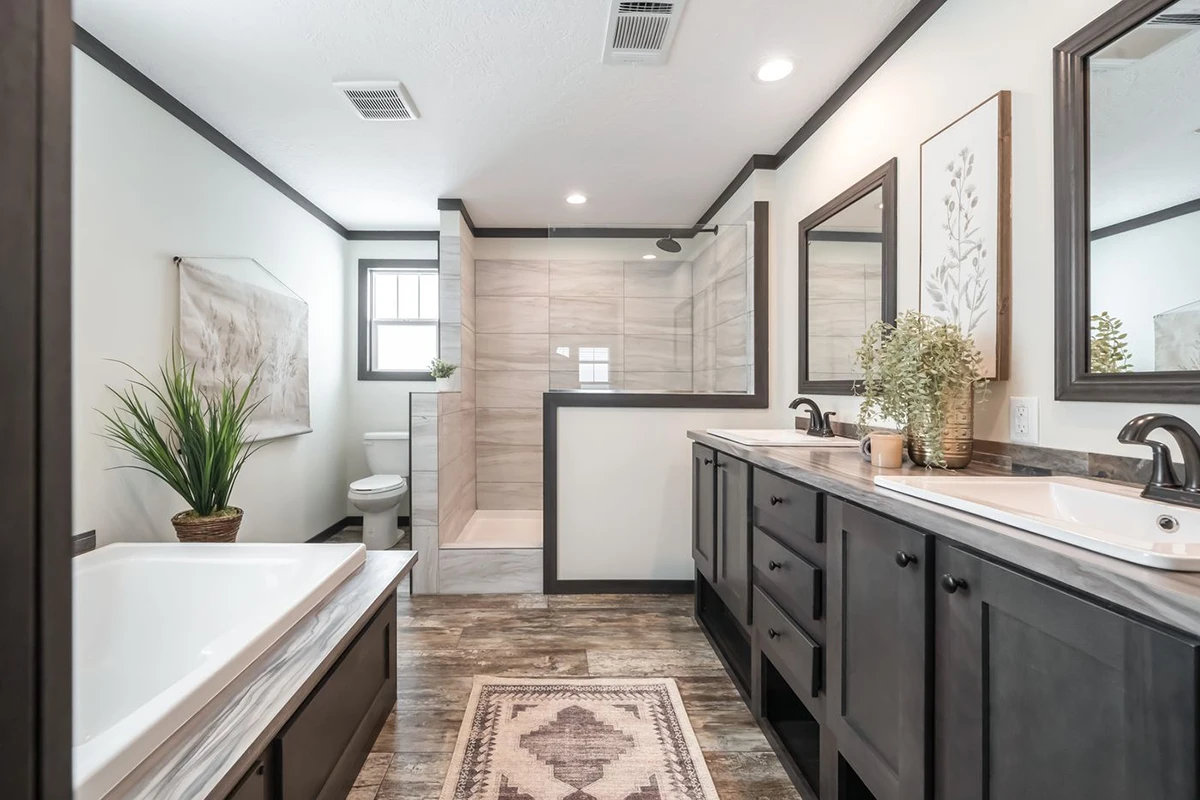 Modern bathroom with double sinks, a bathtub, glass-enclosed shower, toilet, large mirrors, potted plants, and a decorative rug on wood-look flooring.