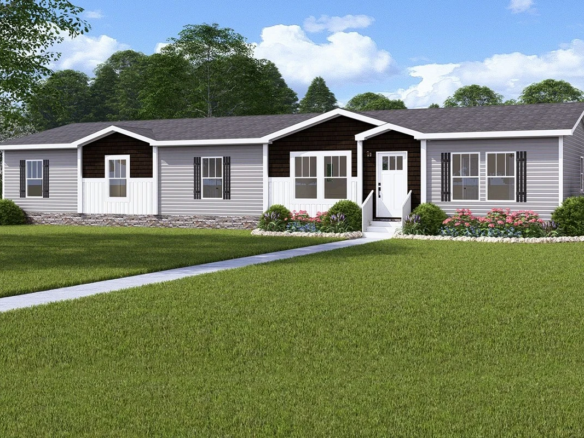 Single-story manufactured home with gray siding, dark shutters, and a front porch, surrounded by landscaped shrubs and a large green lawn. Trees and blue sky in the background.