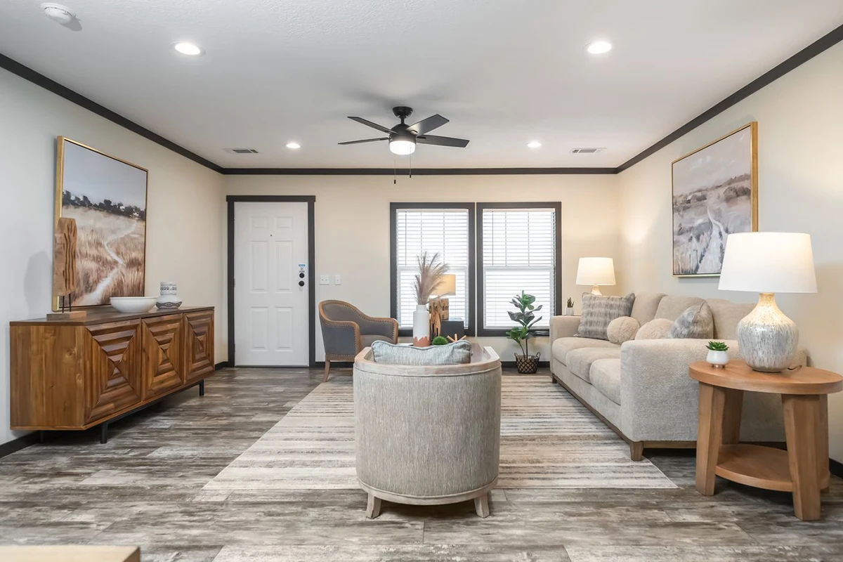 da241167-d40a-4013-bdc3-6826cf6ce2cf A modern living room with neutral decor, light wood furniture, a beige sofa, indoor plants, artwork on the walls, and a ceiling fan. Large windows let in natural light.