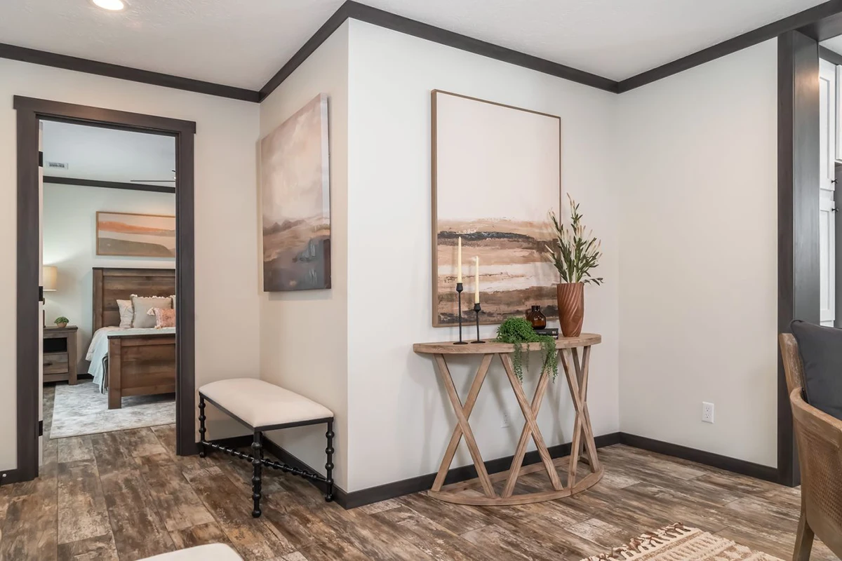 A modern entryway with a round wooden console table, decorative plants, candles, large framed art, and a bench; a bedroom is visible through an open doorway.