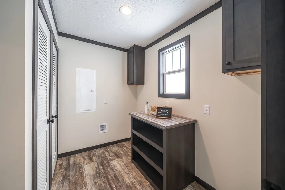 A laundry room with a window, cabinets, open shelving, a countertop, and a small laundry sign on the counter. The floor is wood-patterned vinyl.