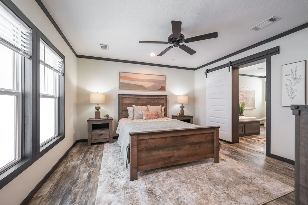Modern bedroom with wooden furniture, two bedside tables with lamps, a ceiling fan, large windows, and a sliding barn door leading to an adjoining bathroom.