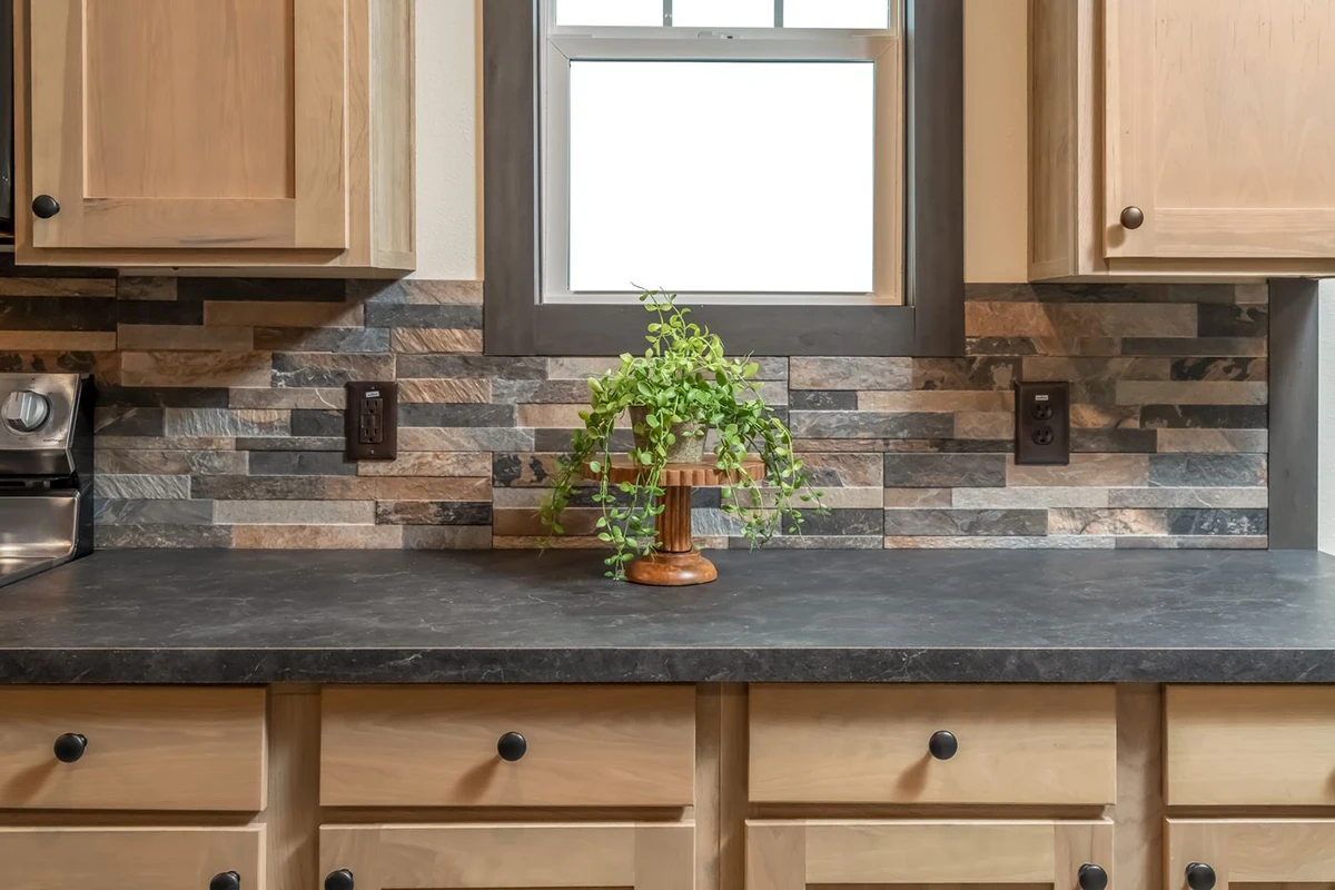 ecd1a0dd-e2d4-440b-afaa-b09b0ac78e11 A potted green plant sits on a dark countertop with light wood cabinets and a multicolored stone tile backsplash beneath a window.