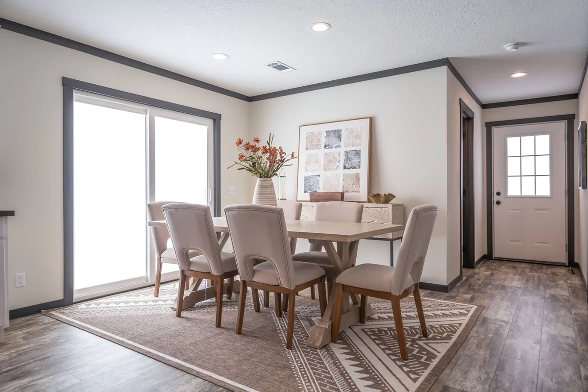 A modern dining room with a light wood table, six upholstered chairs, a geometric rug, wall art, and a vase with flowers near a large sliding glass door.
