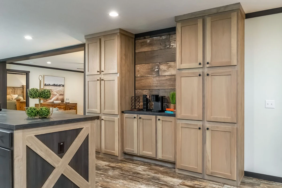 fec00805-f287-44f9-800b-508cc9e26a14 A kitchen with light wood cabinets, a dark countertop, rustic wood accent wall, and coffee maker appliances, featuring wood-look flooring and recessed ceiling lights.