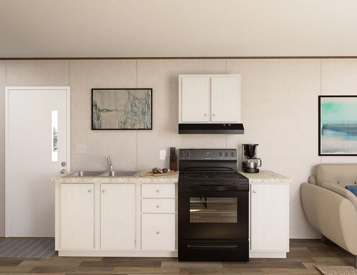 A small kitchen with white cabinets, a black stove and hood, a sink, countertop appliances, and wall art above the sink and next to a beige sofa.