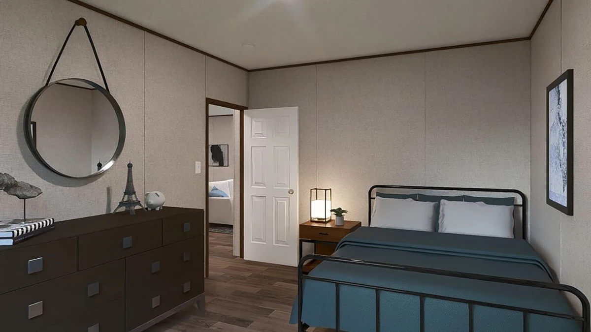 A bedroom with a metal bed frame, blue bedding, nightstand with a lamp, dresser with decor items, round wall mirror, and a door leading to another room.