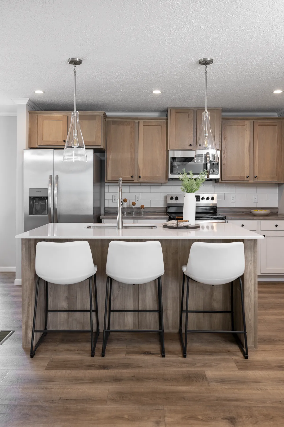 01f626f2-b51e-45e7-b137-bdec1118b9c1 Modern kitchen with wooden cabinets, stainless steel appliances, a central island with a sink, three white bar stools, pendant lights, and a vase with greenery.