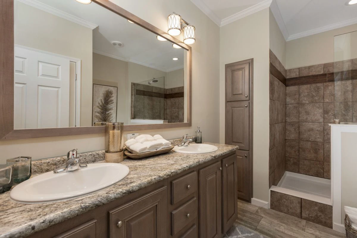 Modern bathroom with double sinks, granite countertop, large mirror, tan cabinets, walk-in shower with brown tiles, and neatly rolled towels on the counter.