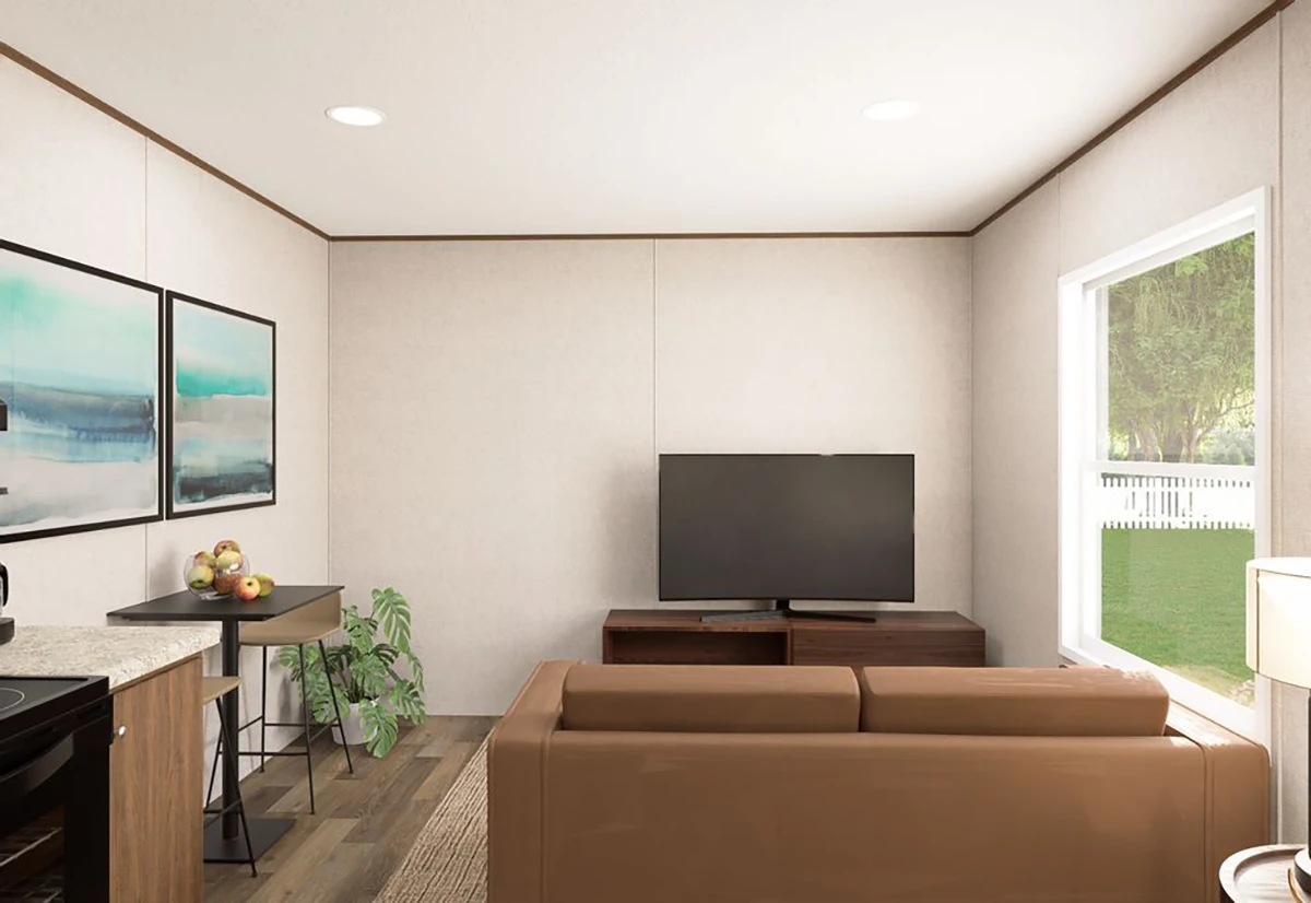A small living room with a brown sofa facing a TV on a stand, large window to the right, and a small kitchen area with bar stools and wall art on the left.