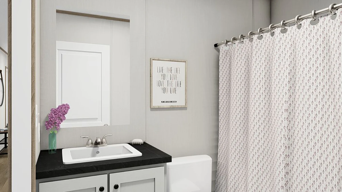 A modern bathroom with a sink, black countertop, vase of purple flowers, wall mirror, framed quote, white toilet, and patterned shower curtain.
