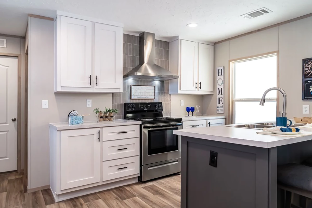Modern kitchen with white cabinets, stainless steel stove and range hood, island with sink, and wood-look flooring. A window and wall decor are visible in the background.