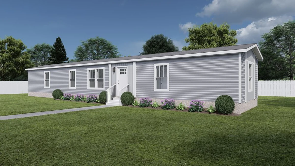 A single-story, light gray manufactured home with white trim, small front porch, and landscaping, situated on a grassy lawn with a white fence and trees in the background.