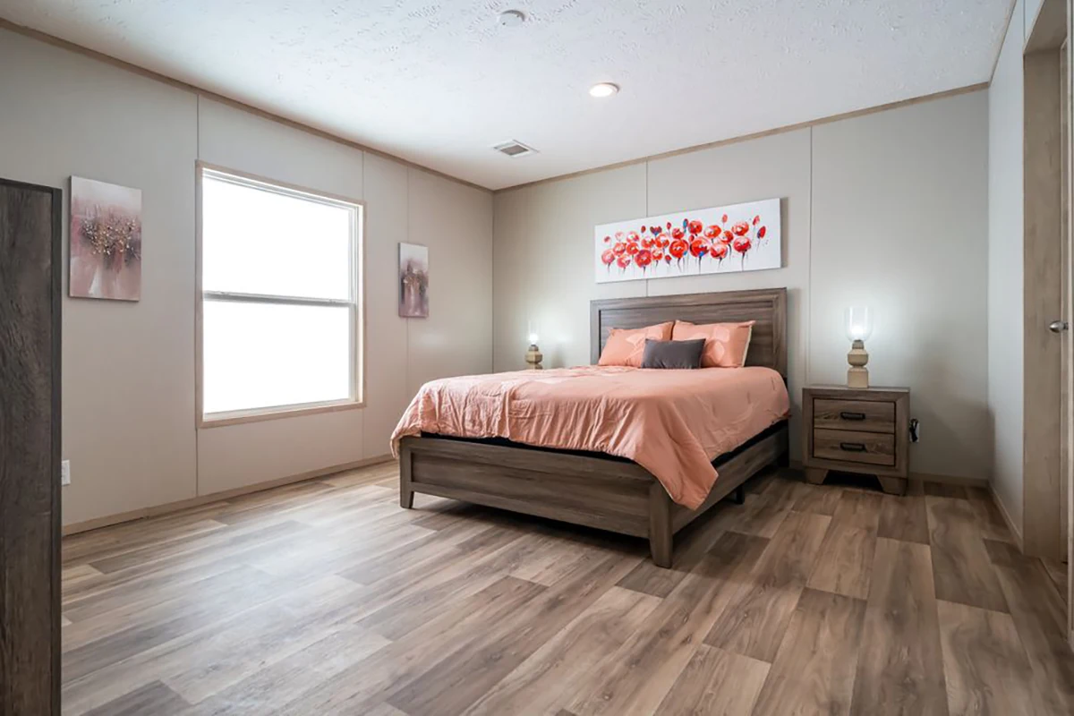 A bedroom with wood flooring, a bed with peach bedding, two nightstands, wall art above the bed, and a large window letting in natural light.