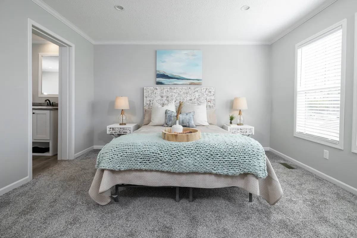 10aea467-7671-451d-9d08-937aa06fac1f Modern bedroom with a neatly made bed, light blue throw blanket, bedside tables with lamps, wall art above the headboard, and sunlight coming through large windows.